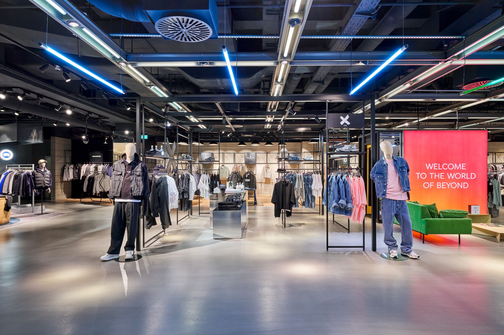 Freistehendes Struktursystem aus schwarzen Metallrahmen mit Kleidung und zwei Schaufensterpuppen in einem modernen Bekleidungsgesch&auml;ft mit sichtbarer Deckeninstallation und gro&szlig;em roten Leuchtschild
