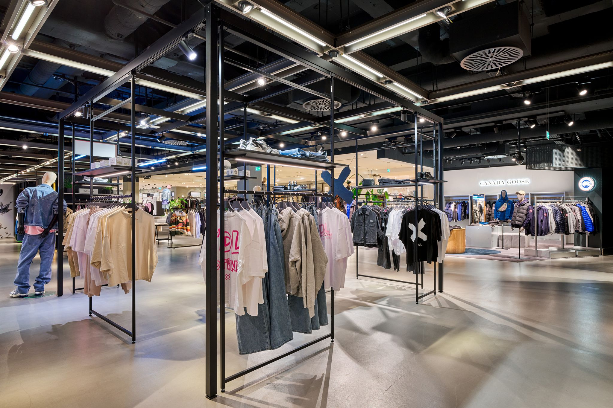 Offener Verkaufsraum mit freistehenden schwarzen Metallgestellen, an denen Kleidung h&auml;ngt, und Mannequins in Jeans und Jacken