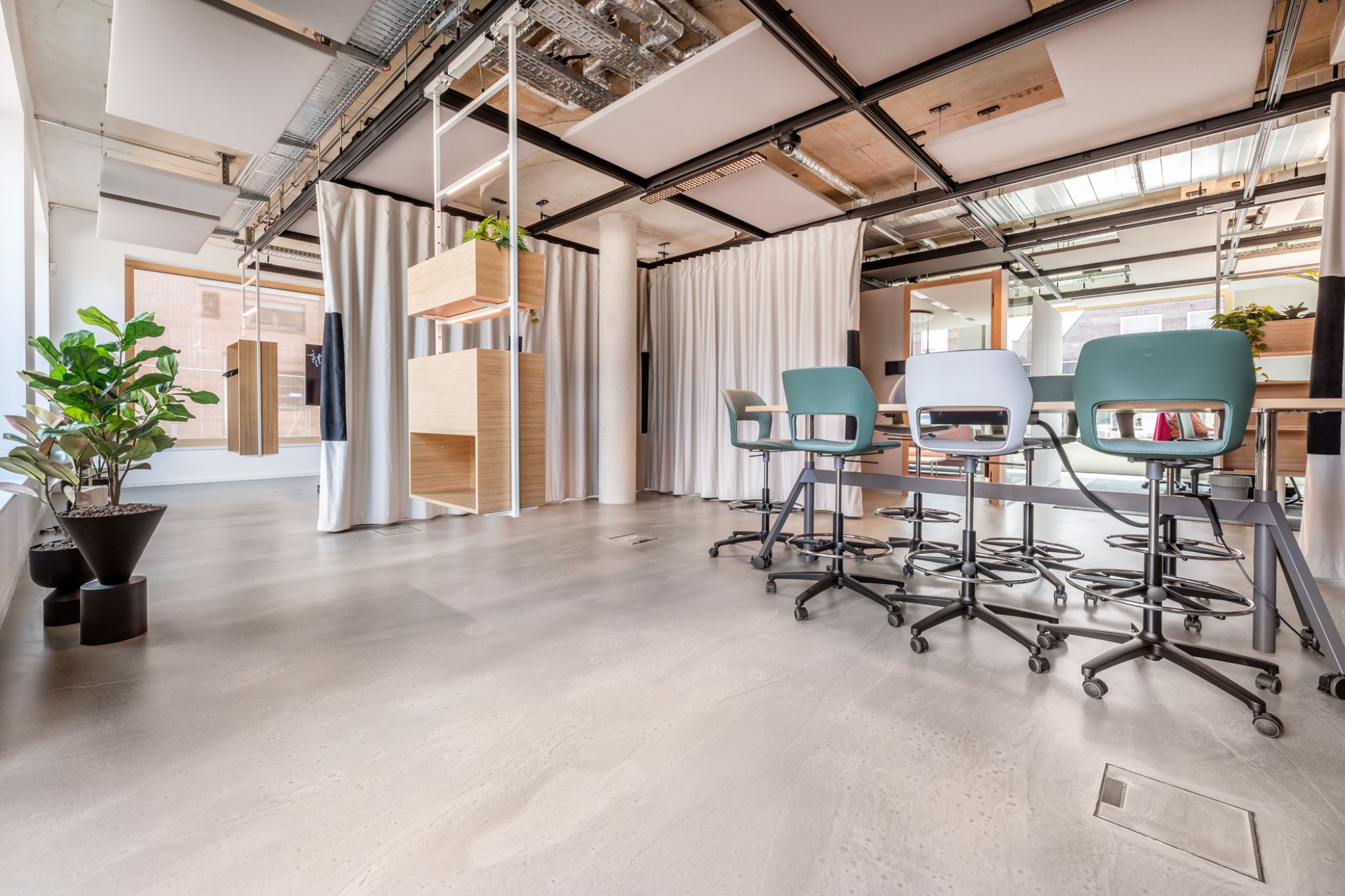 Moderner B&uuml;roraum mit Holz- und Metallm&ouml;beln, grauen Drehst&uuml;hlen, Topfpflanzen und sichtbaren Deckenrohren. Nat&uuml;rliches Licht str&ouml;mt durch gro&szlig;e Fenster und erhellt den Raum.