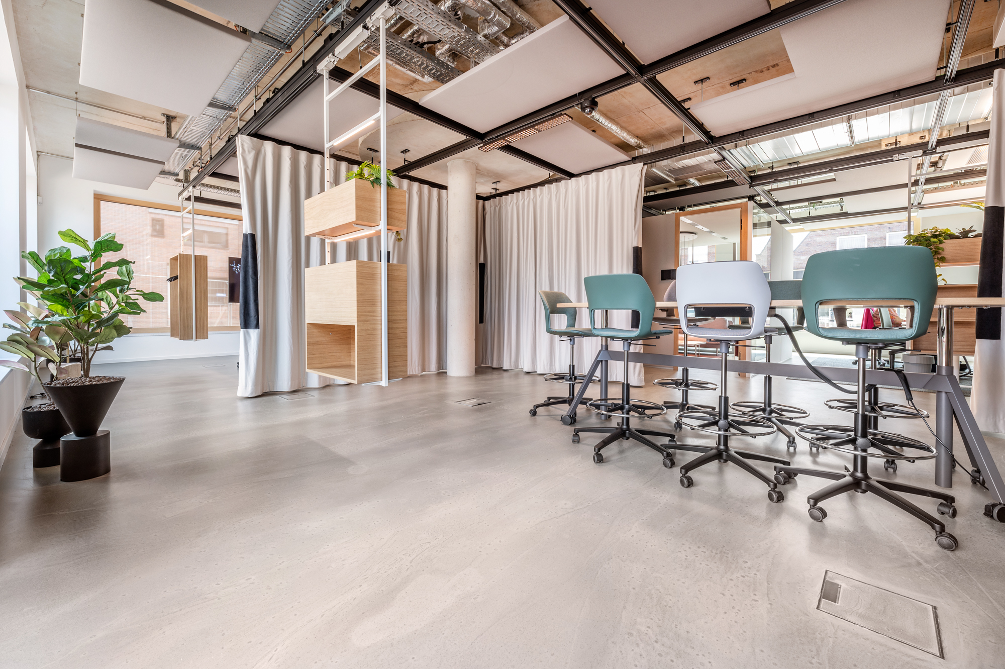 Moderner B&uuml;roraum mit Holz- und Metallm&ouml;beln, grauen Drehst&uuml;hlen, Topfpflanzen und sichtbaren Deckenrohren. Nat&uuml;rliches Licht str&ouml;mt durch gro&szlig;e Fenster und erhellt den Raum.