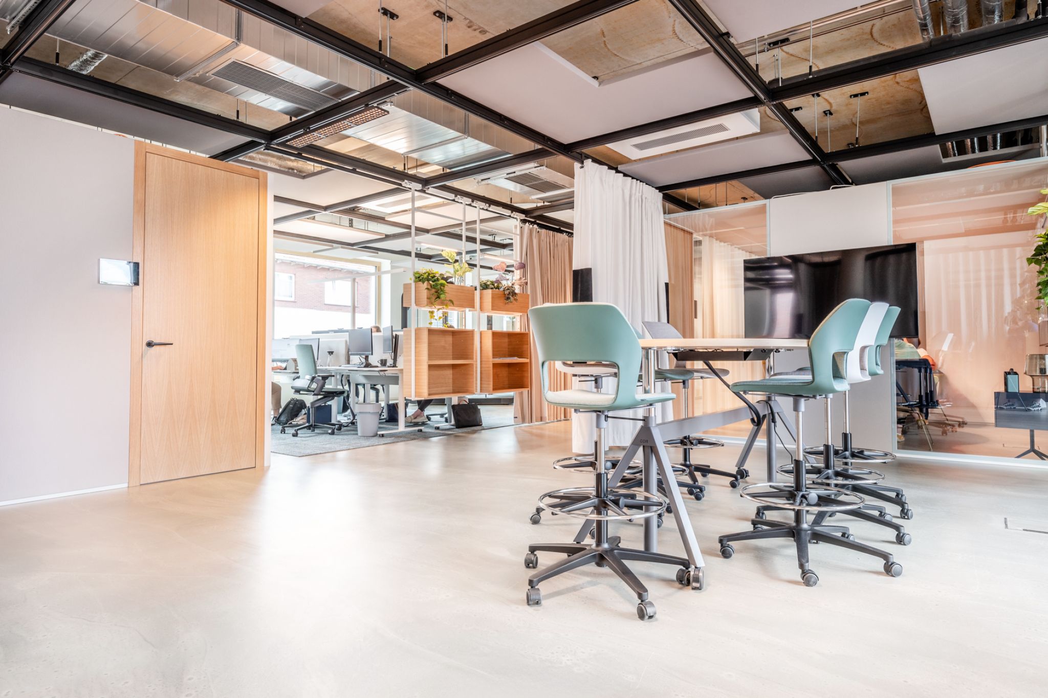 Ein moderner B&uuml;roraum mit hohen St&uuml;hlen um einen Tisch, offenen Arbeitspl&auml;tzen, einer Holzt&uuml;r und nat&uuml;rlichem Licht von den Fenstern.