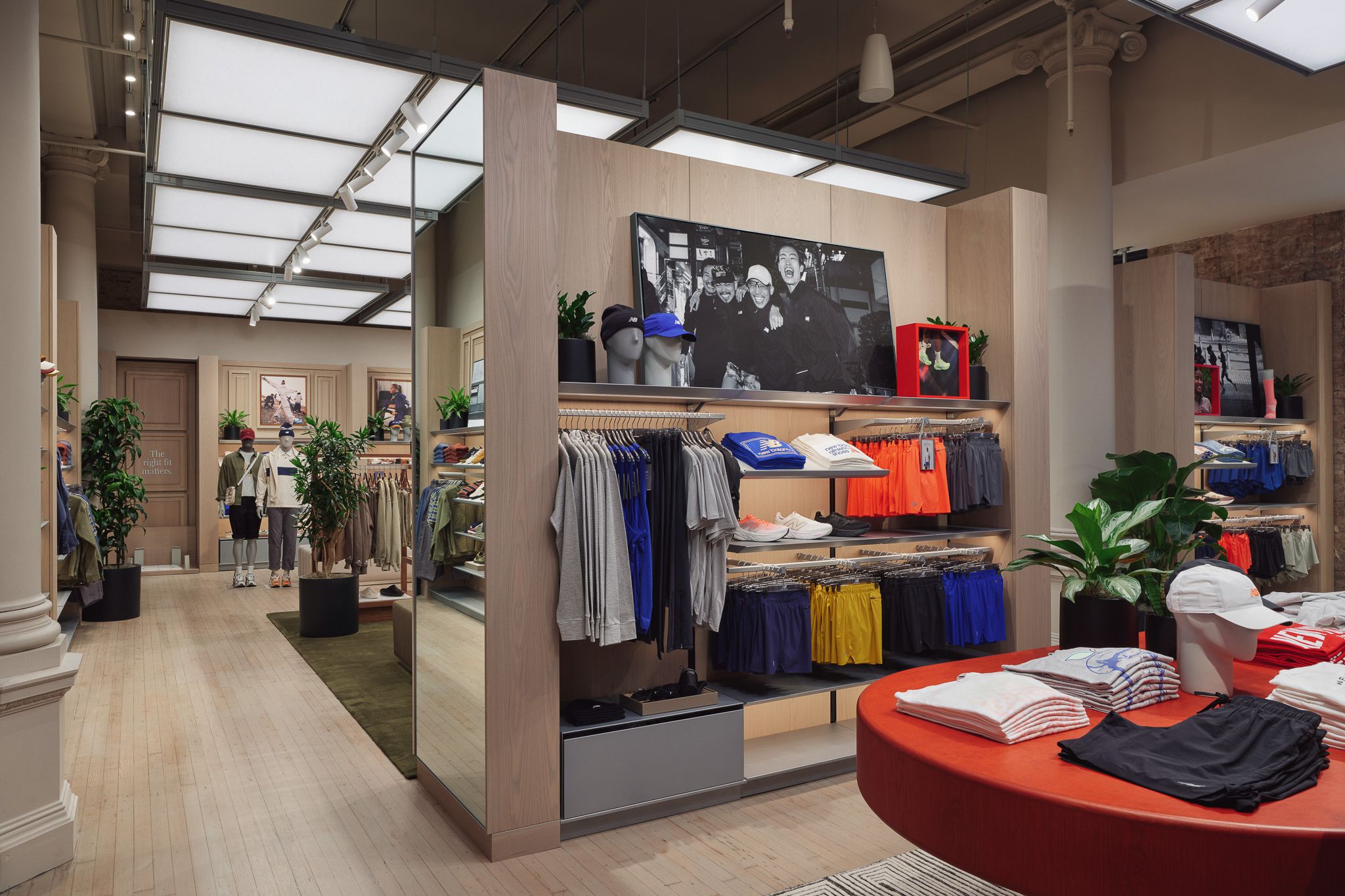 Interior de una tienda de ropa con estantes, mesas, plantas y luces de techo brillantes.