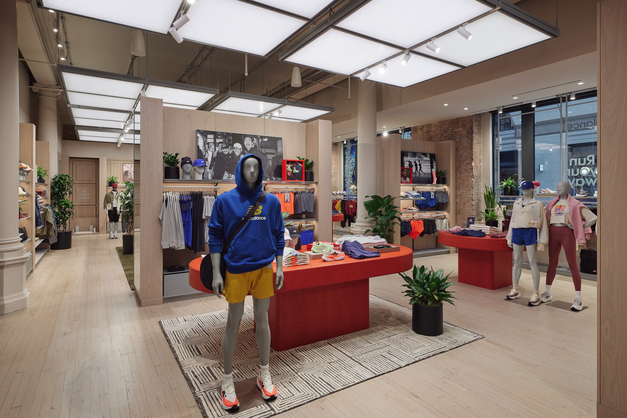 Interior de tienda deportiva con maniqu&iacute;es vestidos con ropa azul y amarilla, mesas de exhibici&oacute;n rojas, plantas y grandes ventanas.