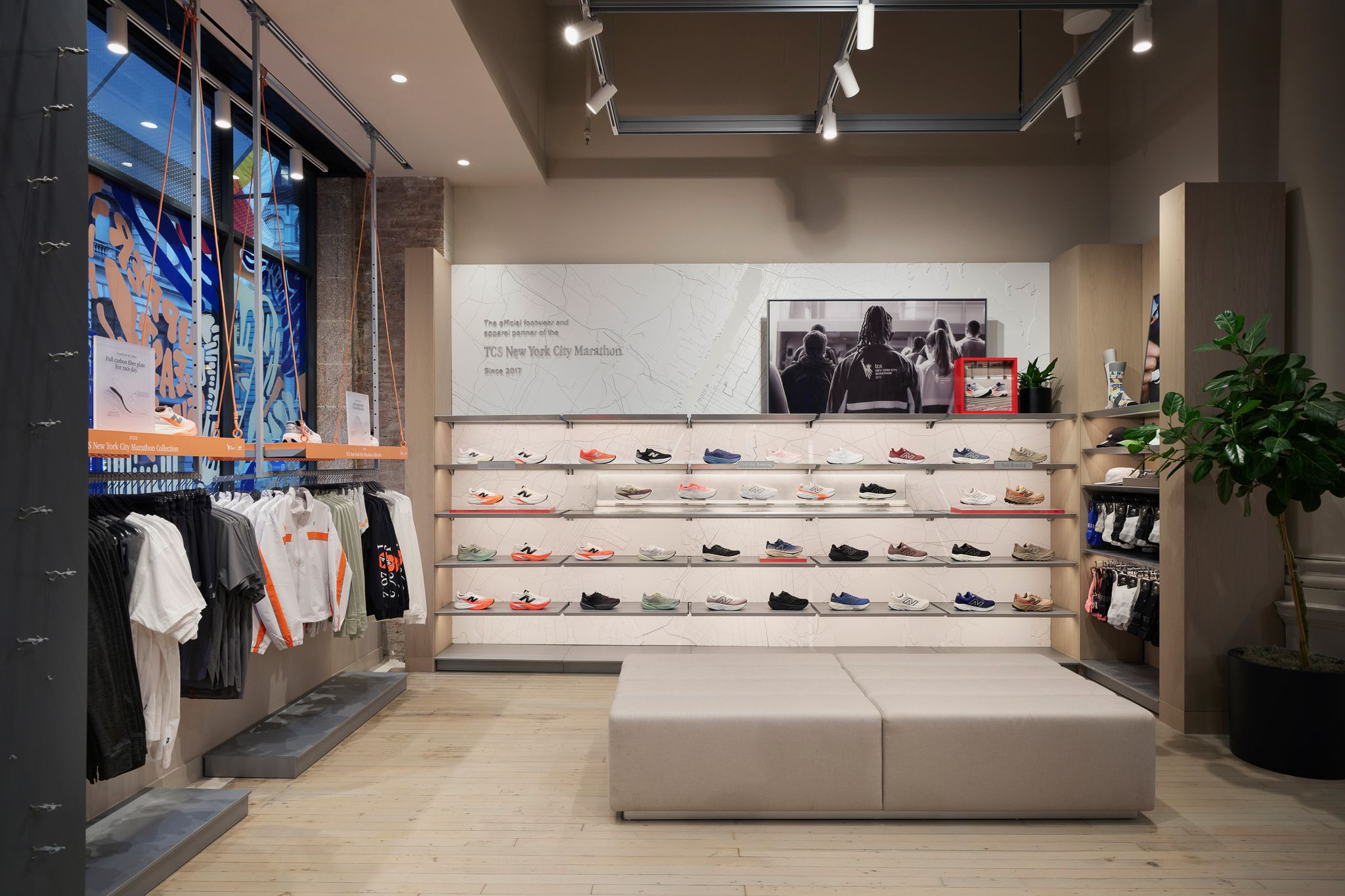 Interior de una tienda de calzado con estantes en la pared que exhiben zapatillas deportivas, un banco central y ropa en la pared izquierda.