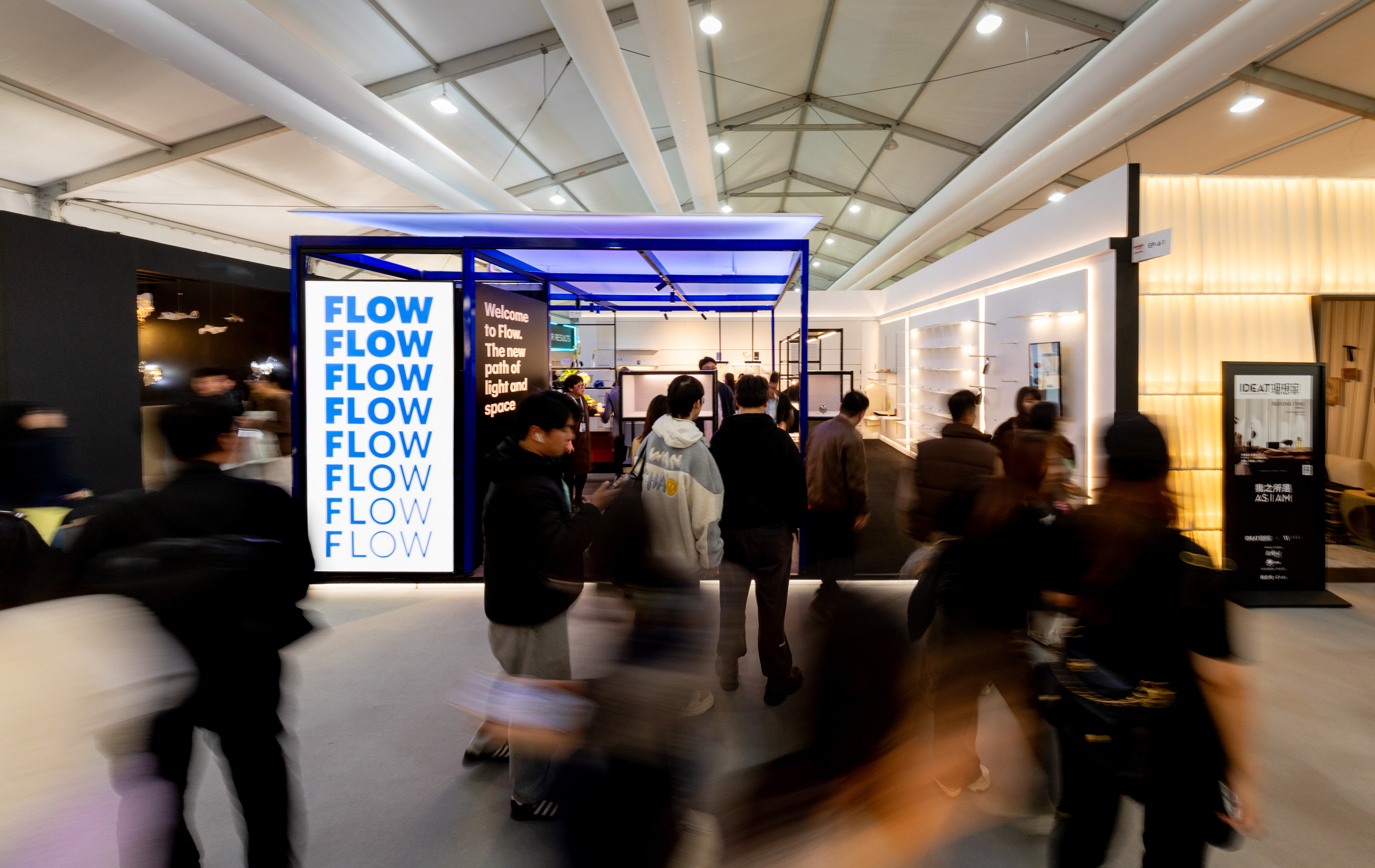 People walking in a modern, brightly lit exhibition hall with a booth displaying the word 'FLOW' in large letters