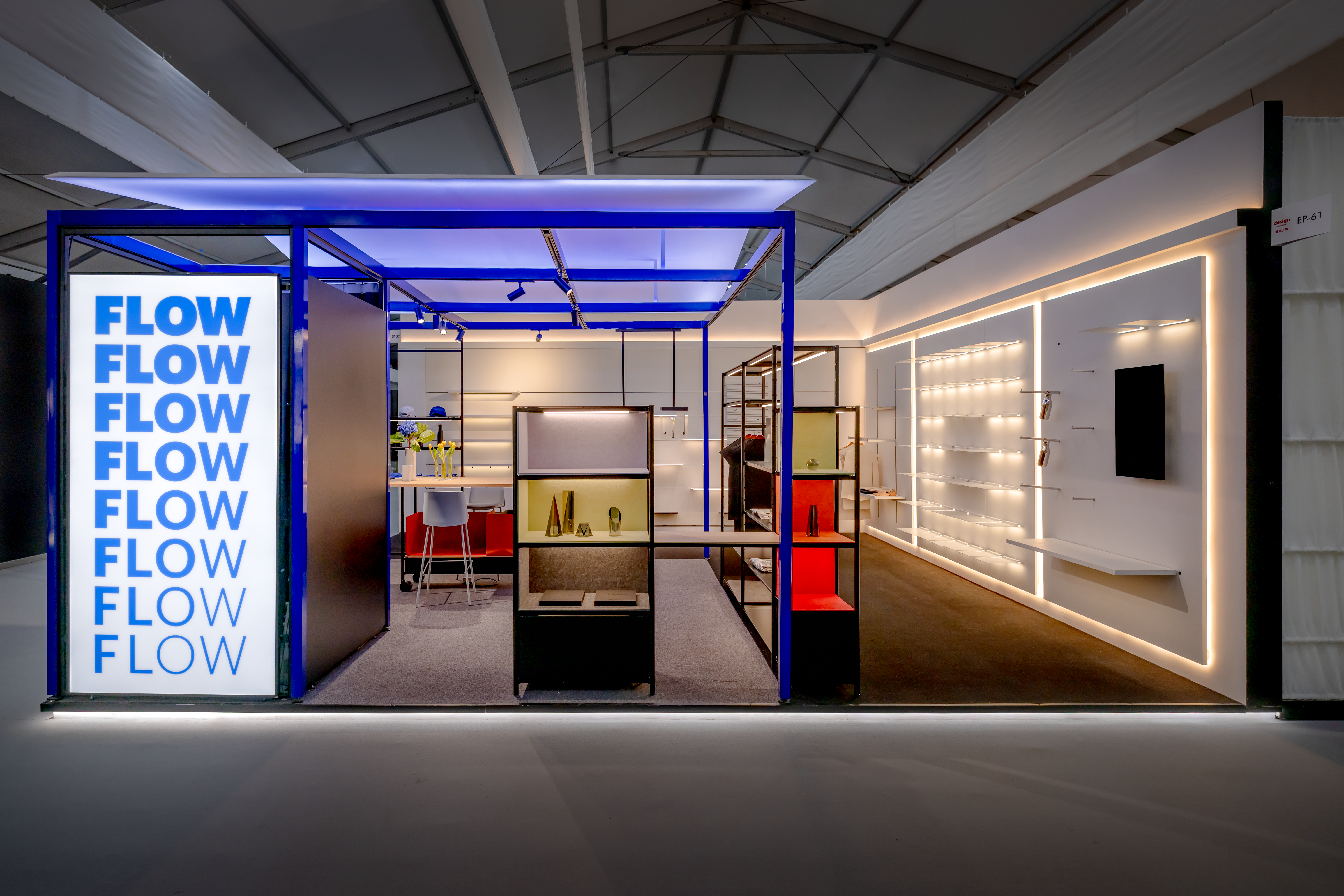 Open exhibition booth with blue metal frame, illuminated 'FLOW' sign, and shelves displaying decorative objects
