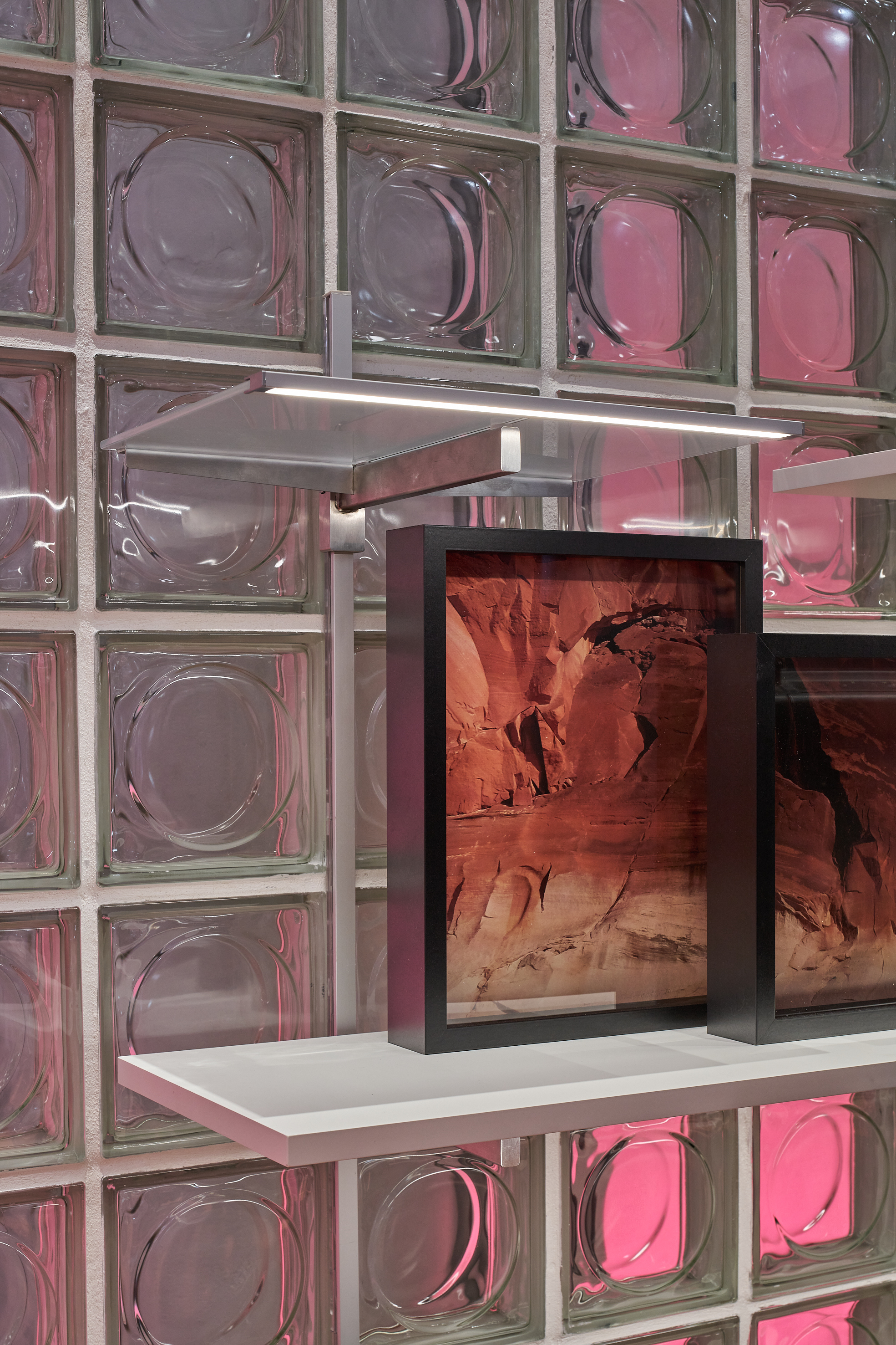 Deux cadres noirs avec des photos de paysage sur une &eacute;tag&egrave;re blanche devant un mur en blocs de verre avec des reflets roses