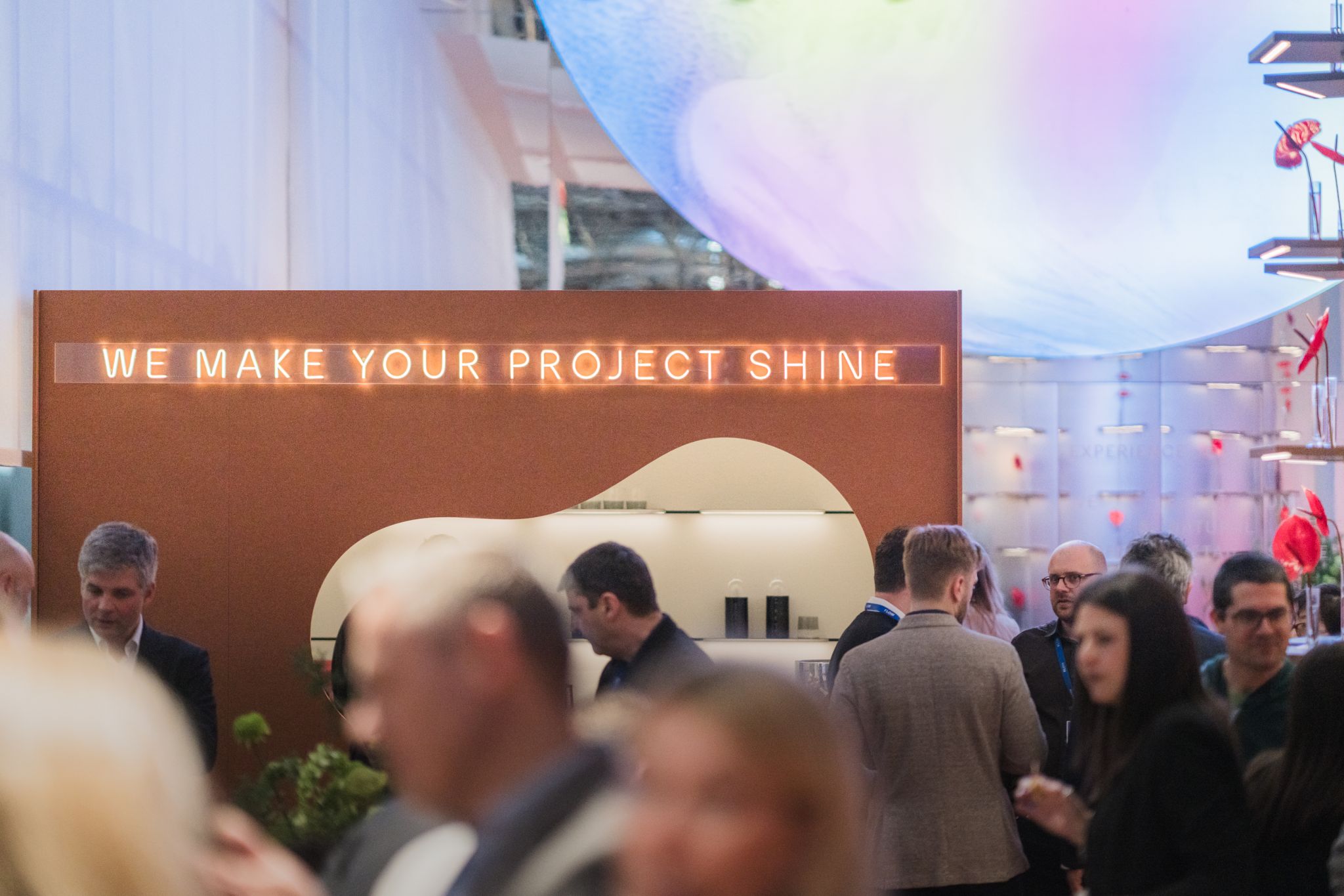 Stand d'exposition avec structure en acier et haut-parleur, enseigne lumineuse 'WE MAKE YOUR PROJECT SHINE' au-dessus, plusieurs visiteurs au premier plan