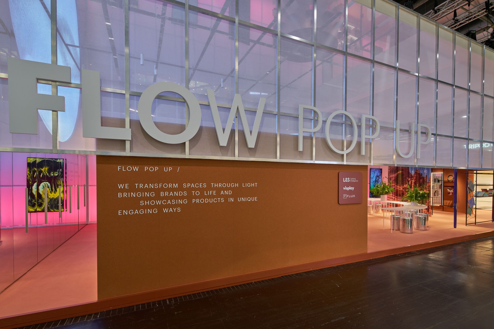Entr&eacute;e d'un stand d'exposition avec grand lettrage &laquo; FLOW POP UP &raquo; et texte sur un mur &agrave; c&ocirc;t&eacute; d'un espace ouvert avec tables et plantes