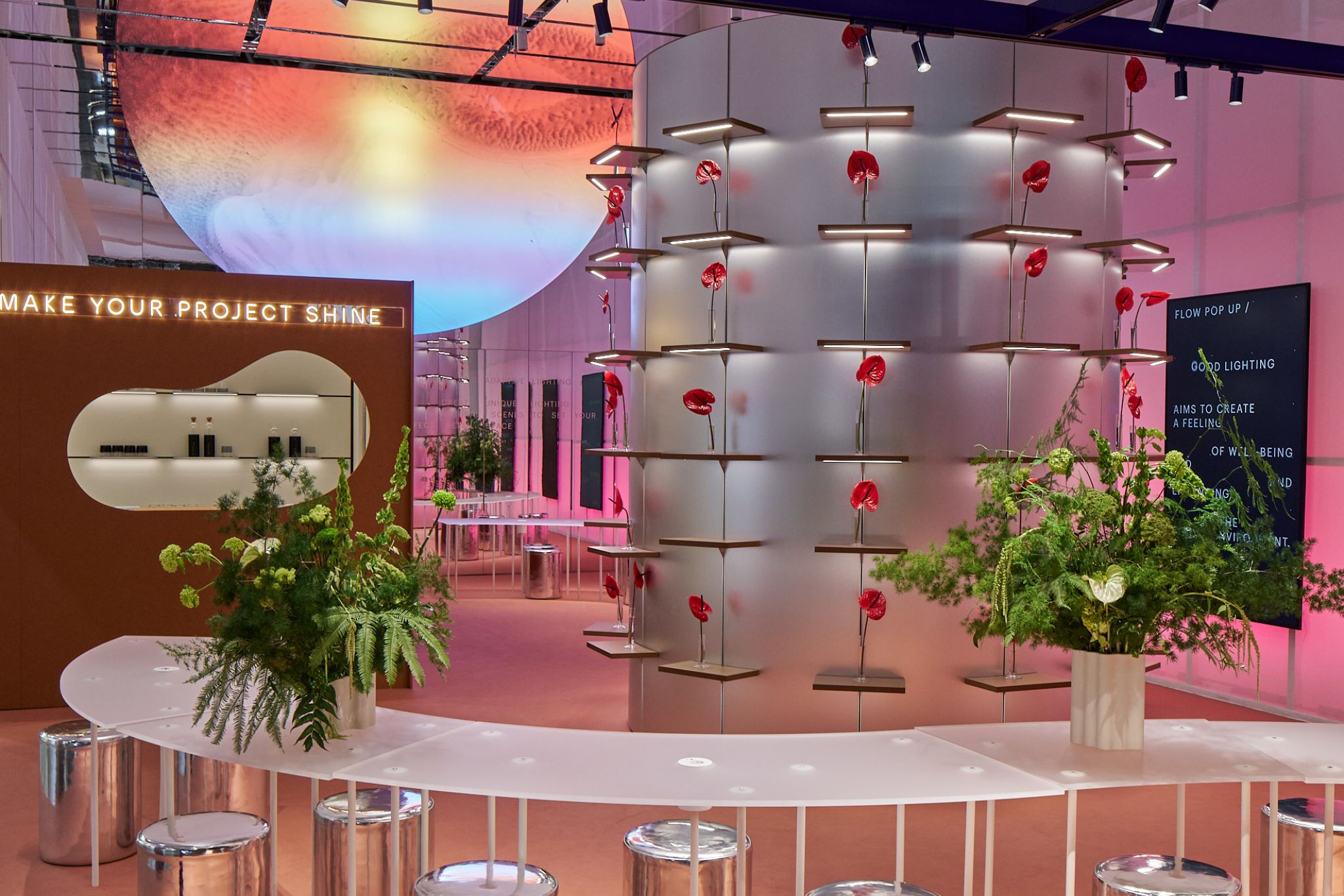 Table blanche incurv&eacute;e avec tabourets argent&eacute;s et plantes vertes au premier plan, colonne ronde avec fleurs rouges et grande sph&egrave;re lumineuse suspendue au plafond en arri&egrave;re-plan