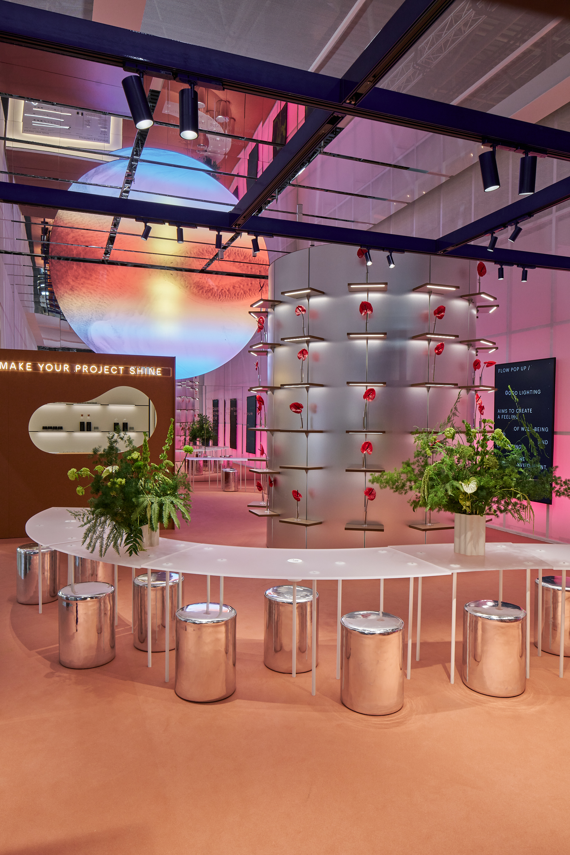 Table blanche incurv&eacute;e avec tabourets argent&eacute;s et plantes vertes au premier plan, colonne ronde avec fleurs rouges et grande sph&egrave;re lumineuse suspendue au plafond en arri&egrave;re-plan