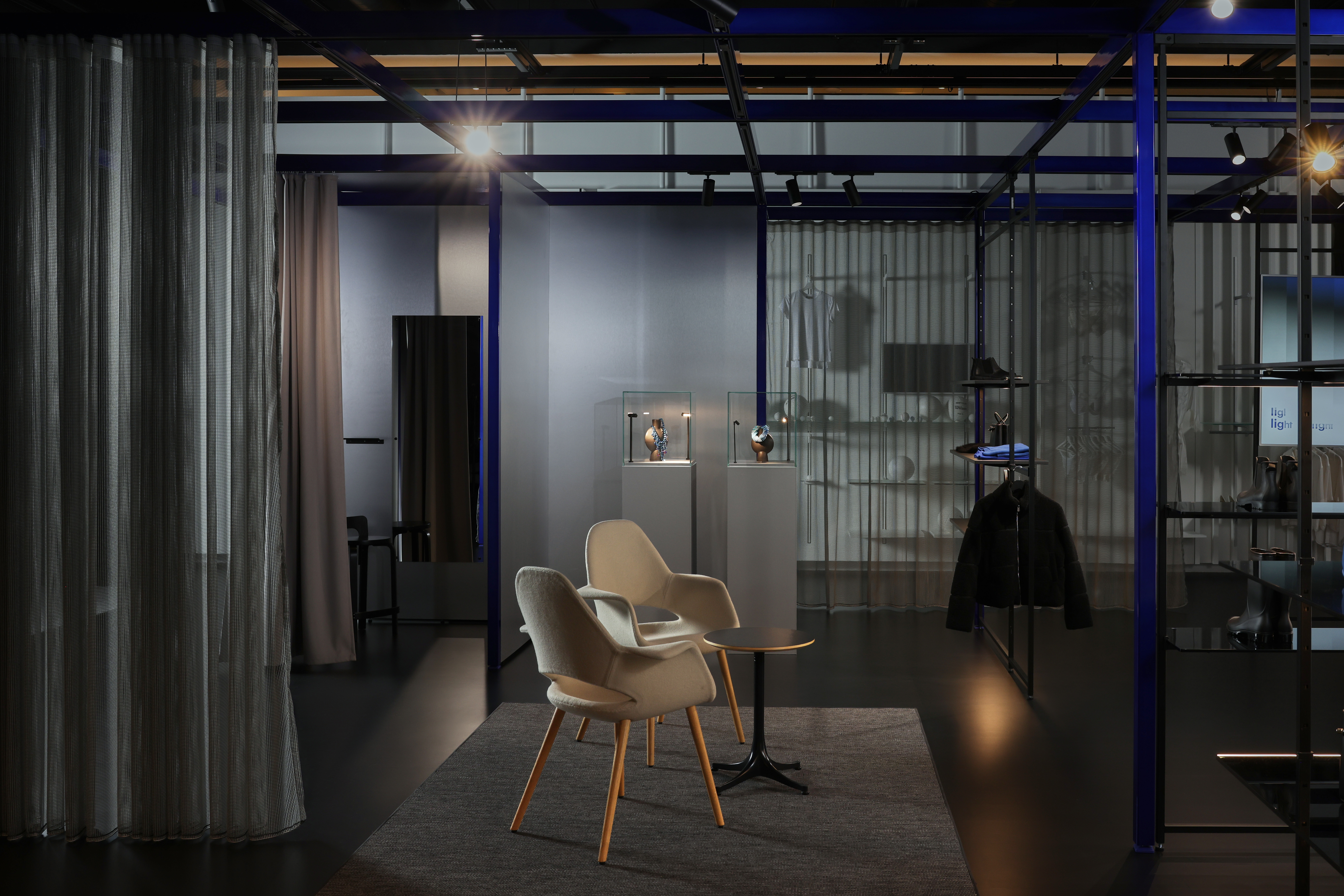 Showroom with black metal shelving system displaying clothing, bags, and accessories, lit and set on a gray carpet.