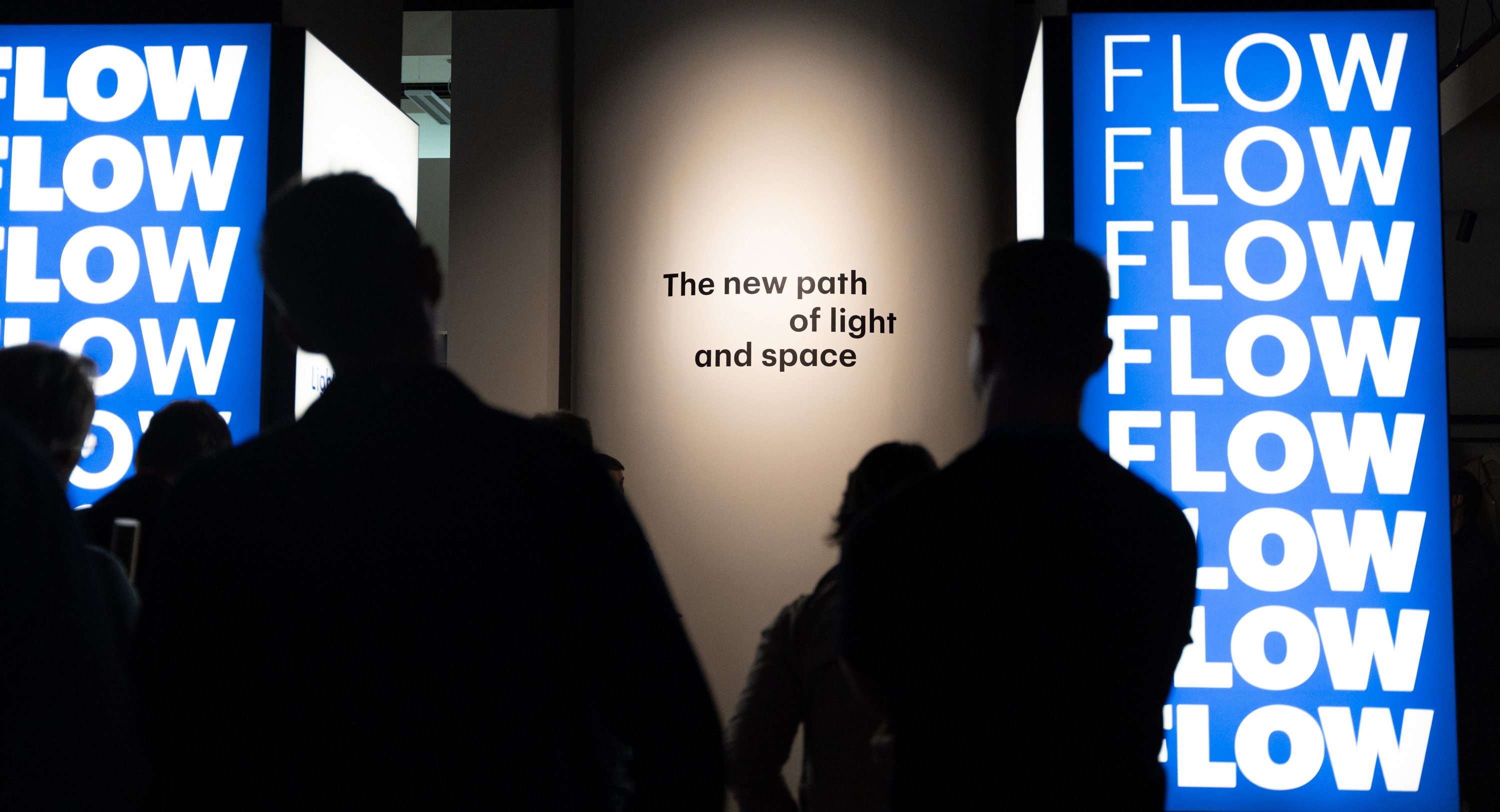 Three silhouetted people in front of a lit wall with the text 'The new path of light and space' and two blue displays showing the word 'FLOW' in white.