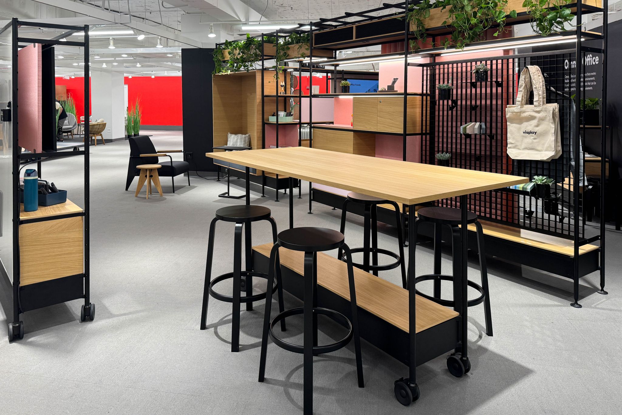 Modern office area with long wooden table on wheels, several black stools, and open shelving with plants and bags.
