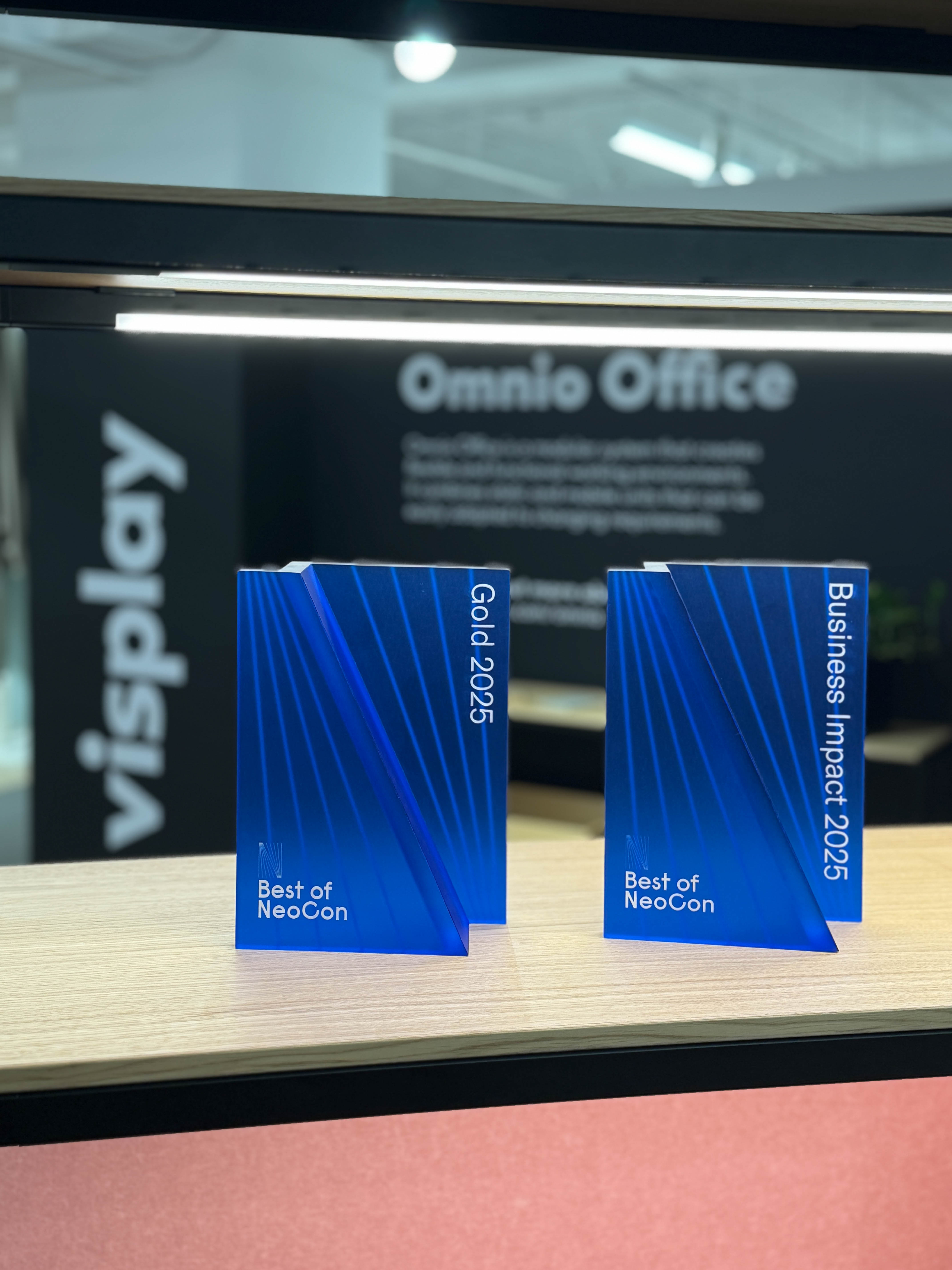 Two blue triangular awards on a light table, labeled 'Gold 2025' and 'Business Impact 2025', each with 'Best of NeoCon'.