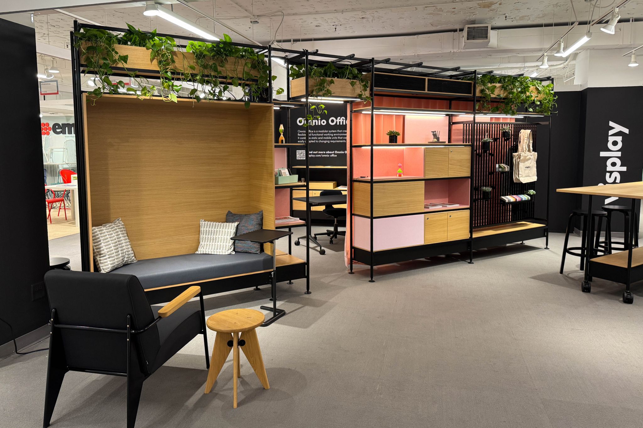 Exhibition booth with modular shelving, seating nook with cushions, black armchair, small wooden stool, and plants on shelves.