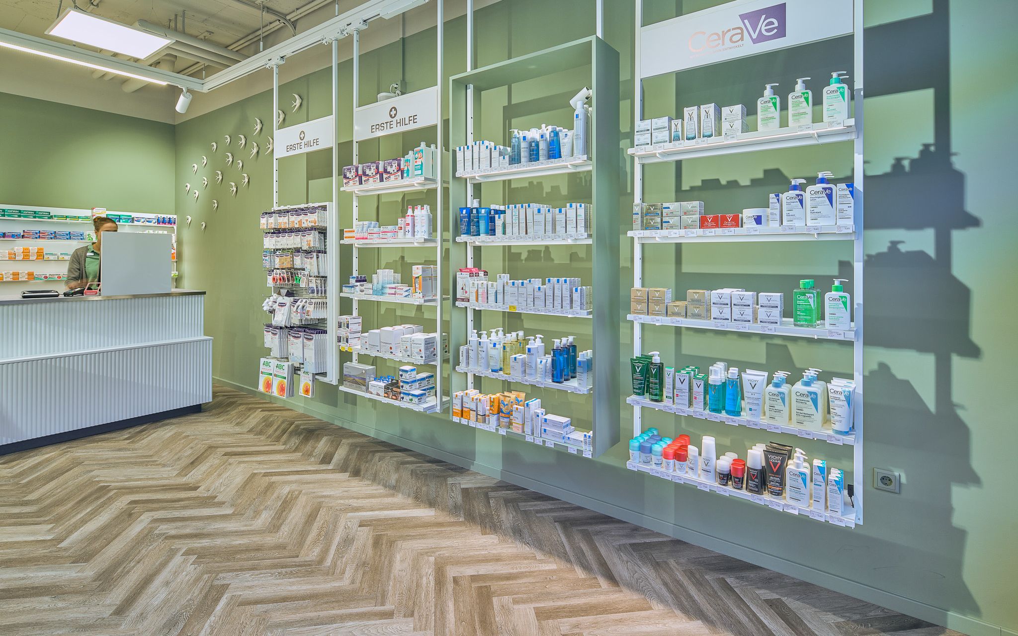 Gr&uuml;ne Wand mit schwebenden Regalen, die verschiedene Arzneimittel und Pflegeprodukte in einer Apotheke pr&auml;sentieren.