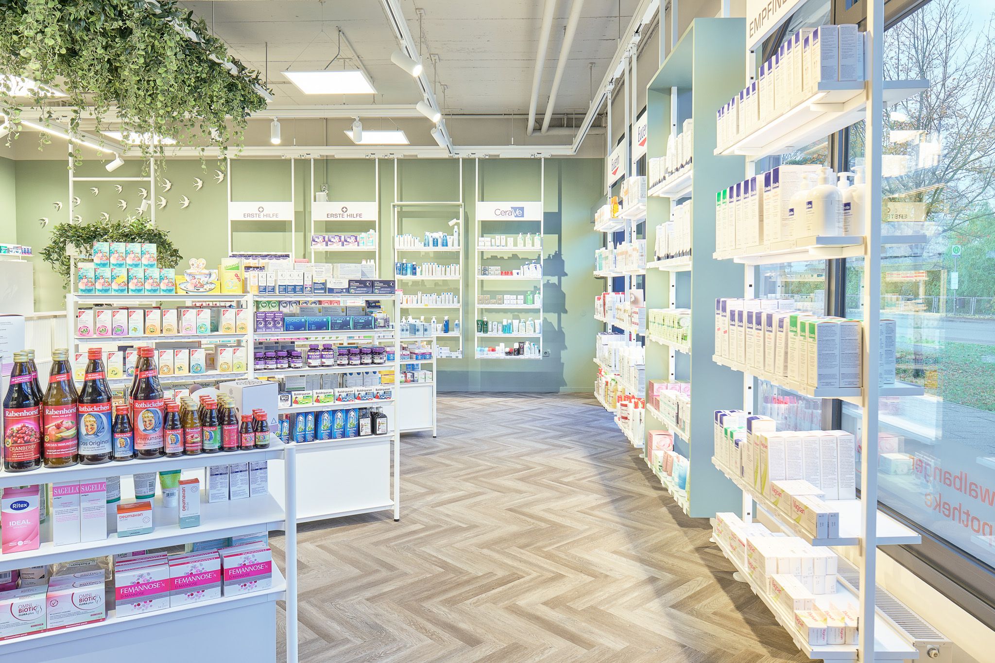 Apothekenregale mit verschiedenen Medikamenten und Produkten, helle Innenraumgestaltung mit gro&szlig;en Fenstern und Pflanzen.