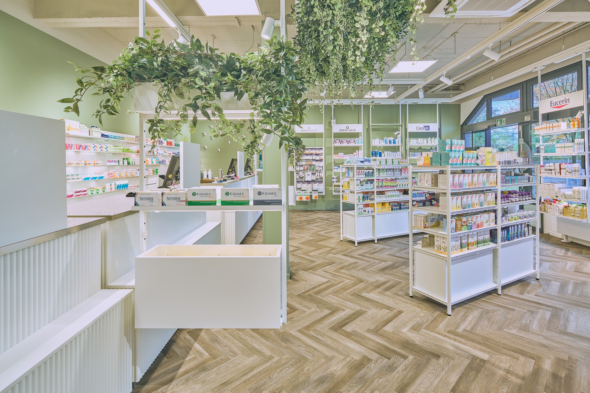 Verkaufsraum einer Apotheke mit wei&szlig;en Regalen, gef&uuml;llt mit Medikamenten und Pflegeprodukten, h&auml;ngenden gr&uuml;nen Pflanzen und Holzboden im Fischgr&auml;tmuster.