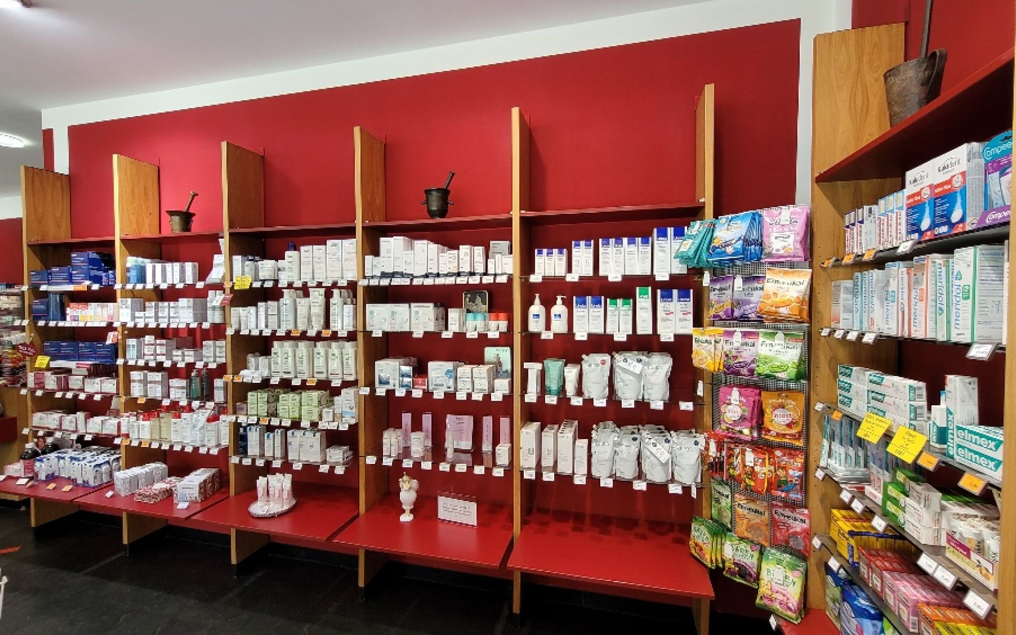 Wandregale mit zahlreichen Medikamentenpackungen und Apothekenartikeln vor roter Wand, rechts h&auml;ngende Packungen und Holzregal mit weiteren Produkten.