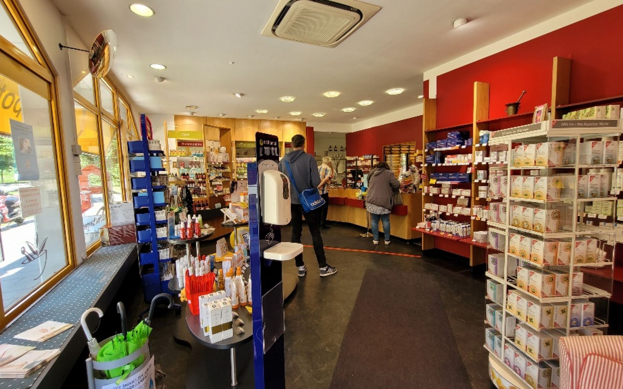 Innenraum einer Apotheke mit roten W&auml;nden, Regalen voller Produkte und zwei Kunden am Tresen.