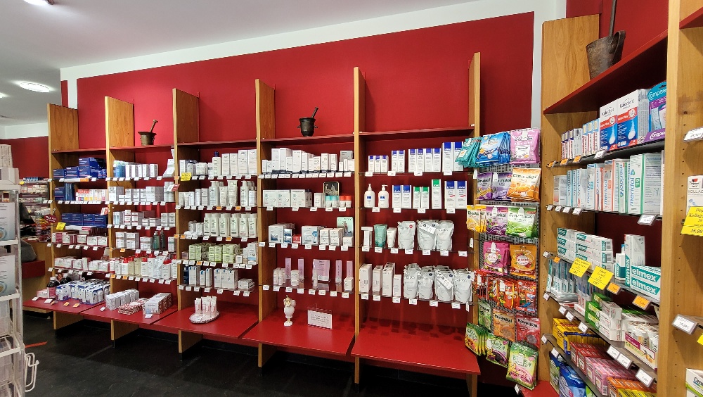 Estantes de pared con numerosas cajas de medicamentos y art&iacute;culos de farmacia frente a una pared roja, paquetes colgados y estante de madera con m&aacute;s productos a la derecha.