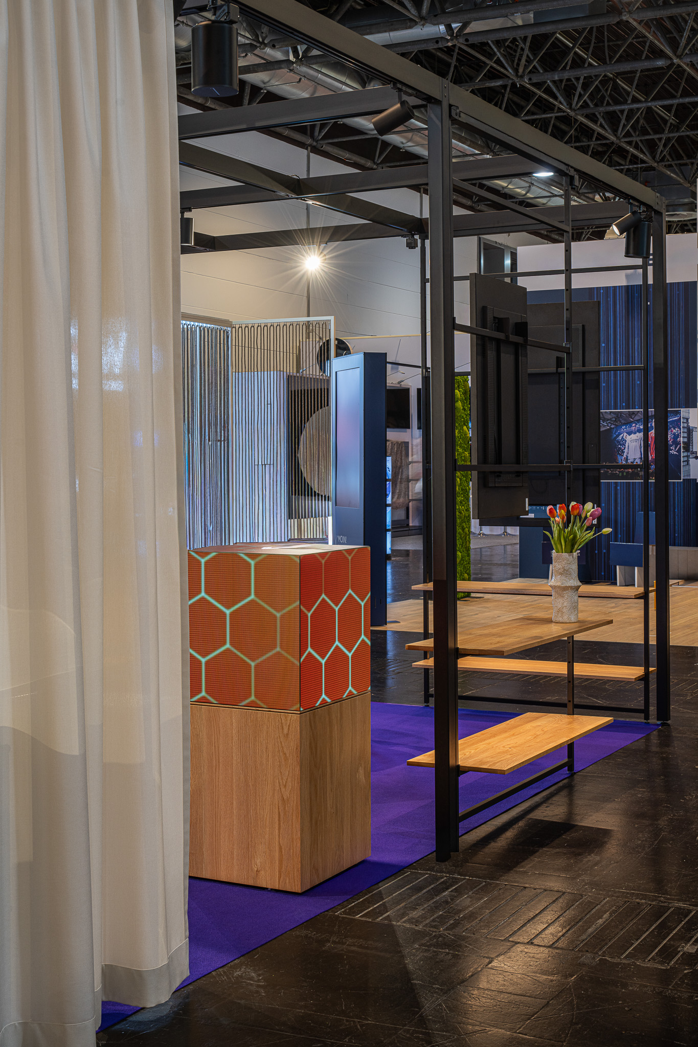 Messestand mit schwarzem Metallrahmen, Holzregalen, orange-hexagonaler Dekoration und Vase mit Tulpen auf blauem Boden