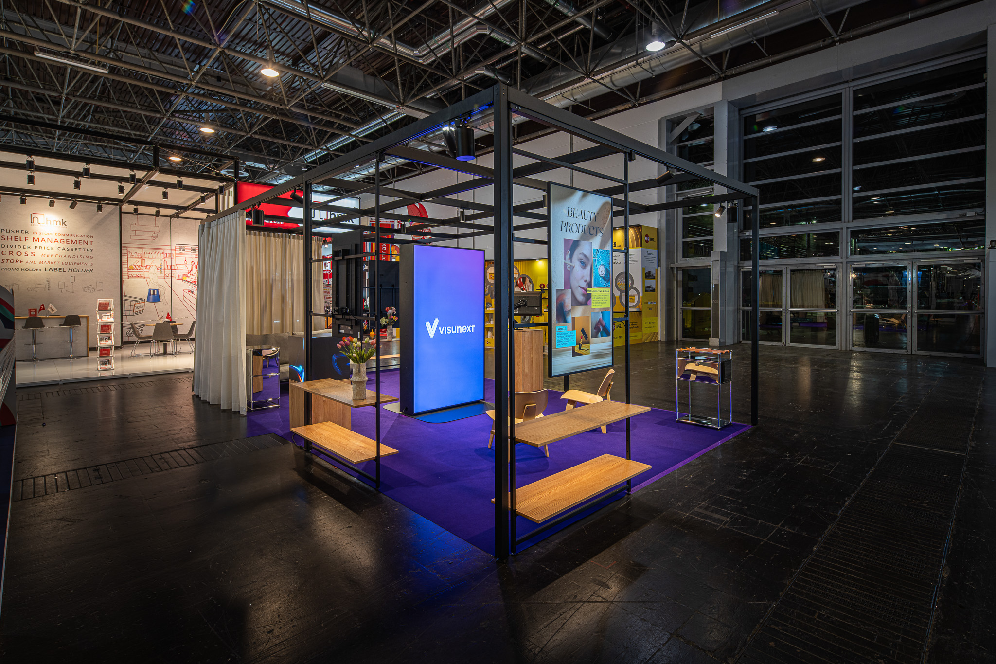Offener Messestand mit schwarzem Metallrahmen, Holzsitzb&auml;nken, violettem Teppich und zwei gro&szlig;en Bildschirmen, einer zeigt das Logo 'visunext'
