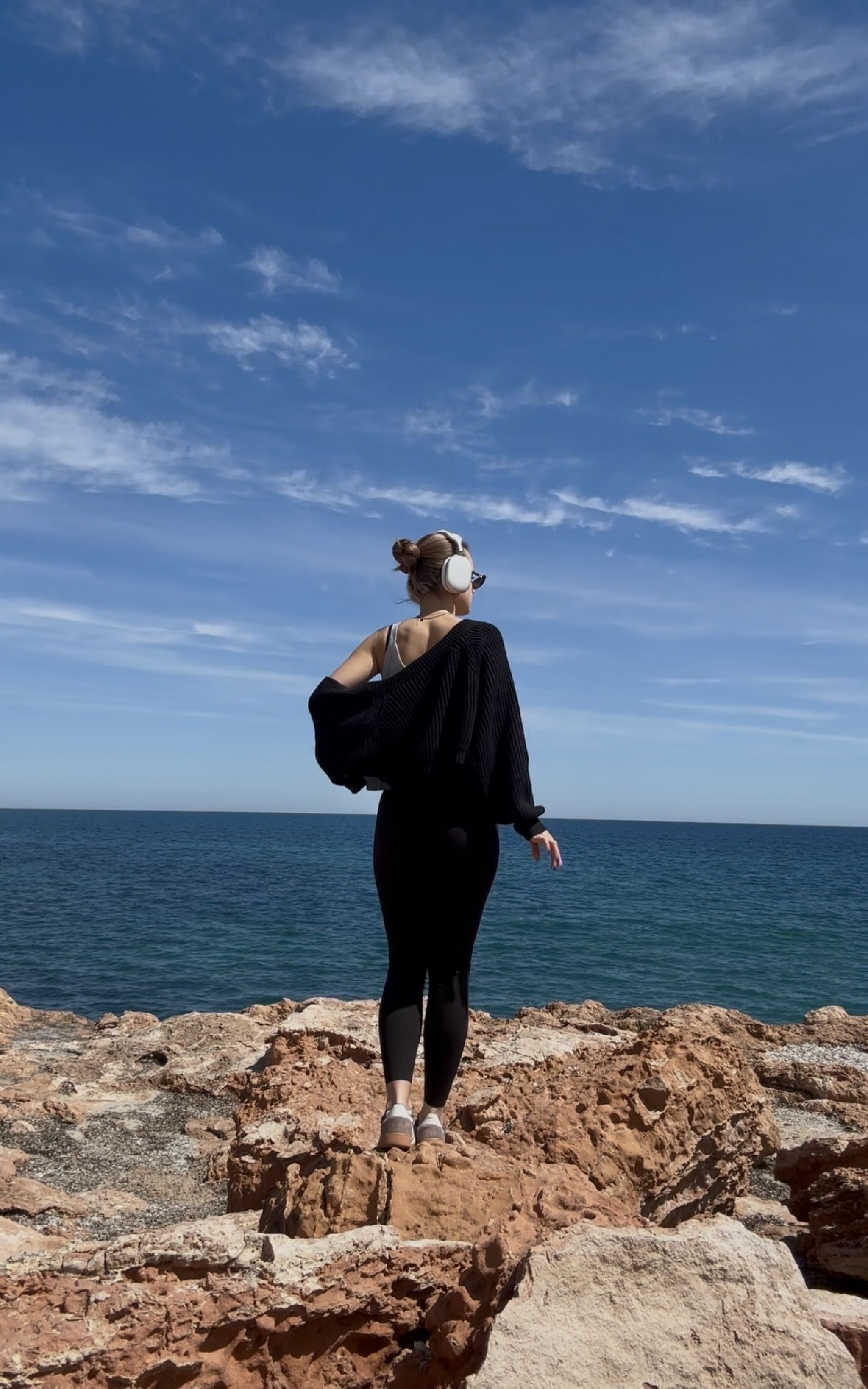 Frau mit Kopfh&ouml;rern steht auf felsiger K&uuml;ste und blickt aufs Meer unter blauem Himmel.