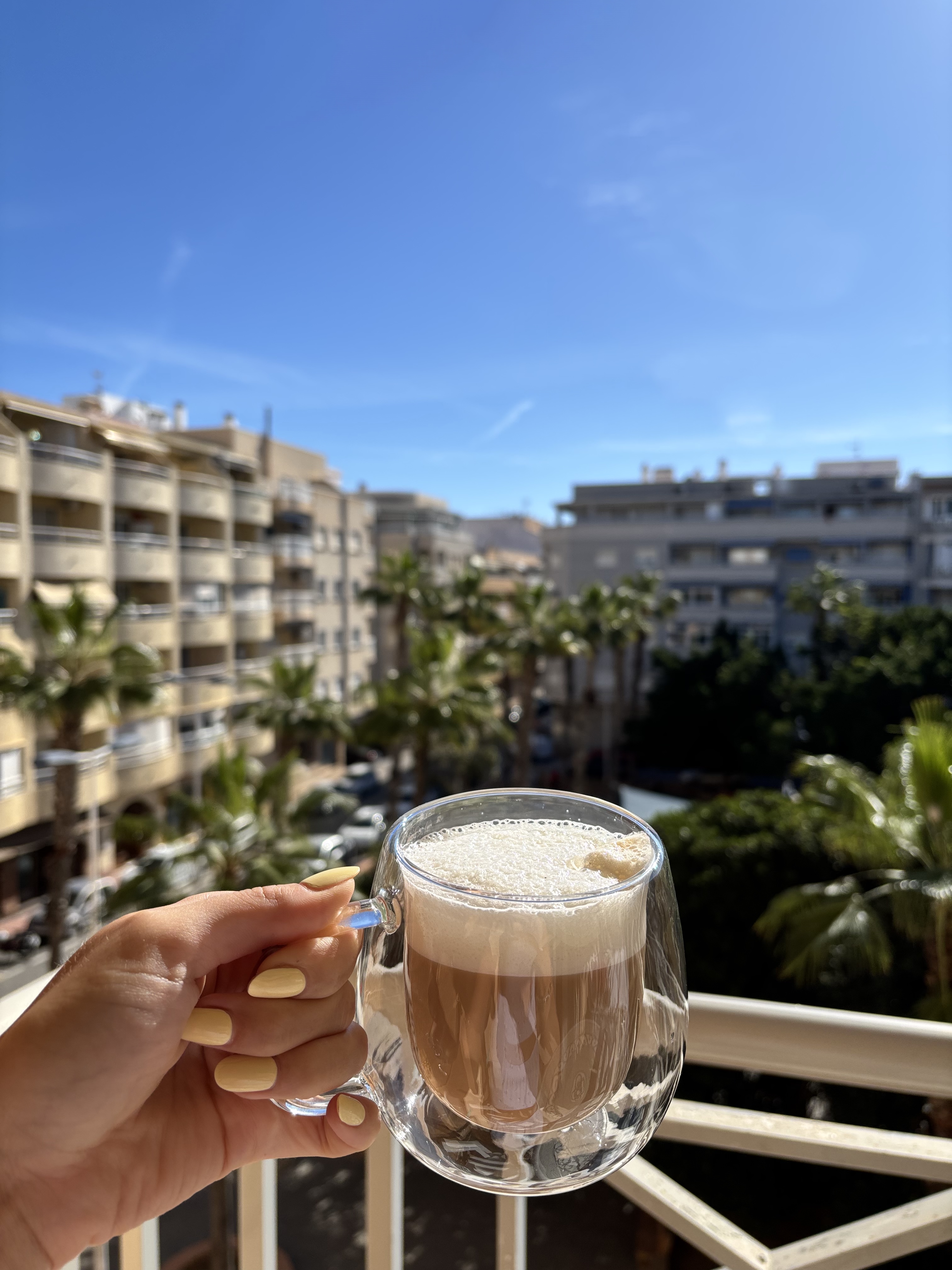 Hand mit gelb lackierten Fingern&auml;geln h&auml;lt eine Tasse Cappuccino mit Milchschaum vor einer Kulisse aus Palmen und Wohngeb&auml;uden.