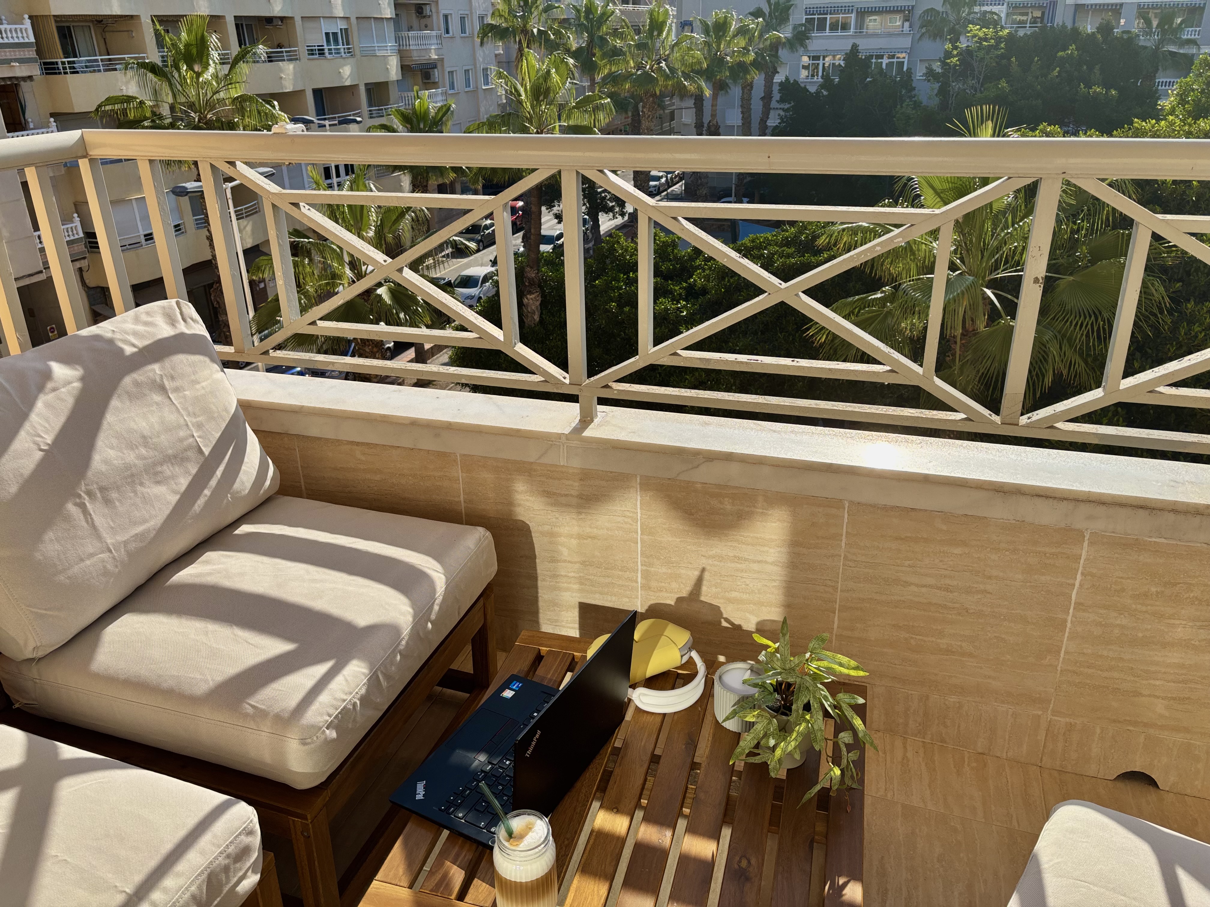 Balkon mit zwei gepolsterten St&uuml;hlen, Holztisch mit Laptop, Kerze und Pflanze, Blick auf Palmen und Wohngeb&auml;ude.